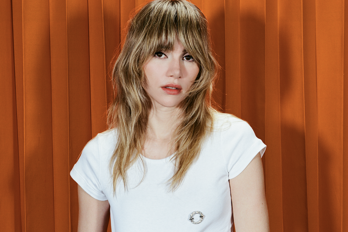 Singer and actress Suki Waterhouse wearing a white t-shirt with blonde shaggy bangs against an orange backdrop.